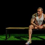 A muscular, tattooed man seated on a bench in a gym, depicting modern fitness lifestyle.