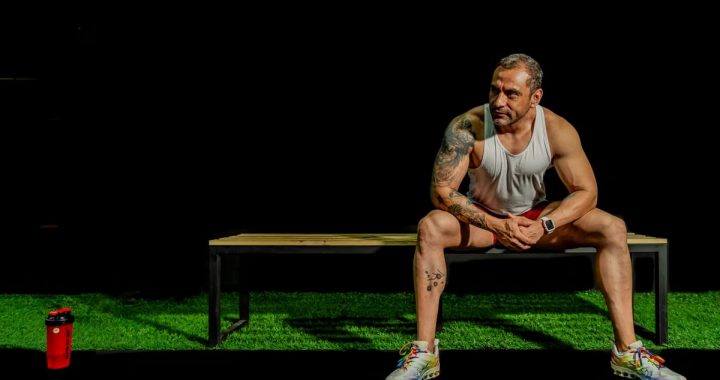 A muscular, tattooed man seated on a bench in a gym, depicting modern fitness lifestyle.
